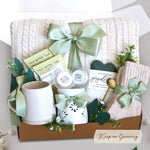 The Keep on Growing Gift Box xome with a cream cable knit blanket tied with a sage ribbon, Wildflower Pop Beehive Mug, Starburst wax warmer and 2 wax melts, 2 tea bags, Seaside Soak bath salts, cozy boot socks, and gold demitasse spoon