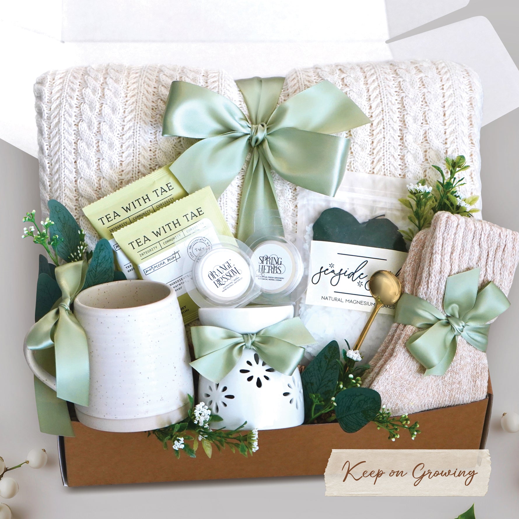 The Keep on Growing Gift Box xome with a cream cable knit blanket tied with a sage ribbon, Wildflower Pop Beehive Mug, Starburst wax warmer and 2 wax melts, 2 tea bags, Seaside Soak bath salts, cozy boot socks, and gold demitasse spoon