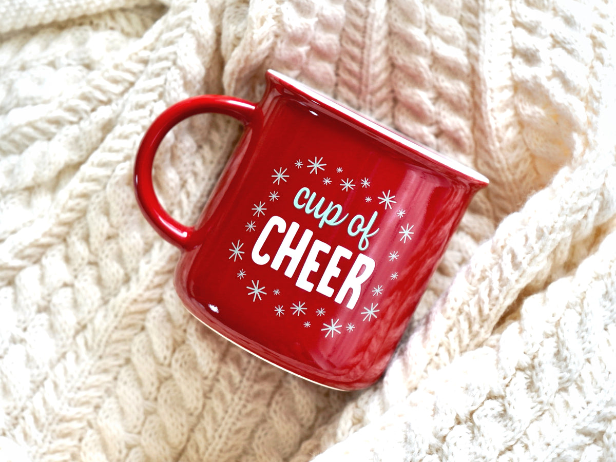 Holiday mug shown is a red ceramic mug with a white rim with the words "cup of cheer" surrounded by snowflakes 