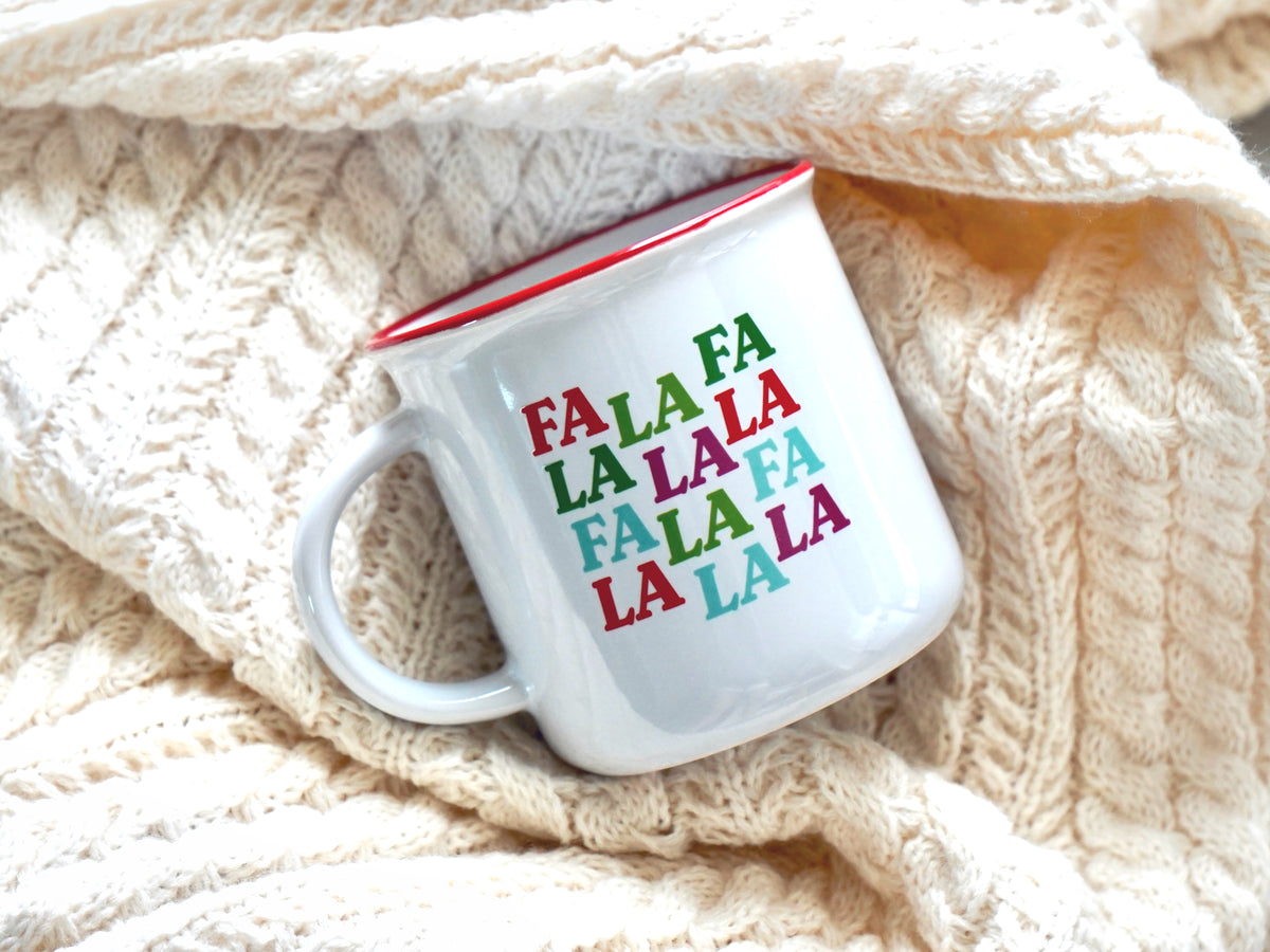 Holiday mug shown is a white ceramic mug with a red rim with the words Fa La Fa La La La Fa La Fa La La La in multi colors 