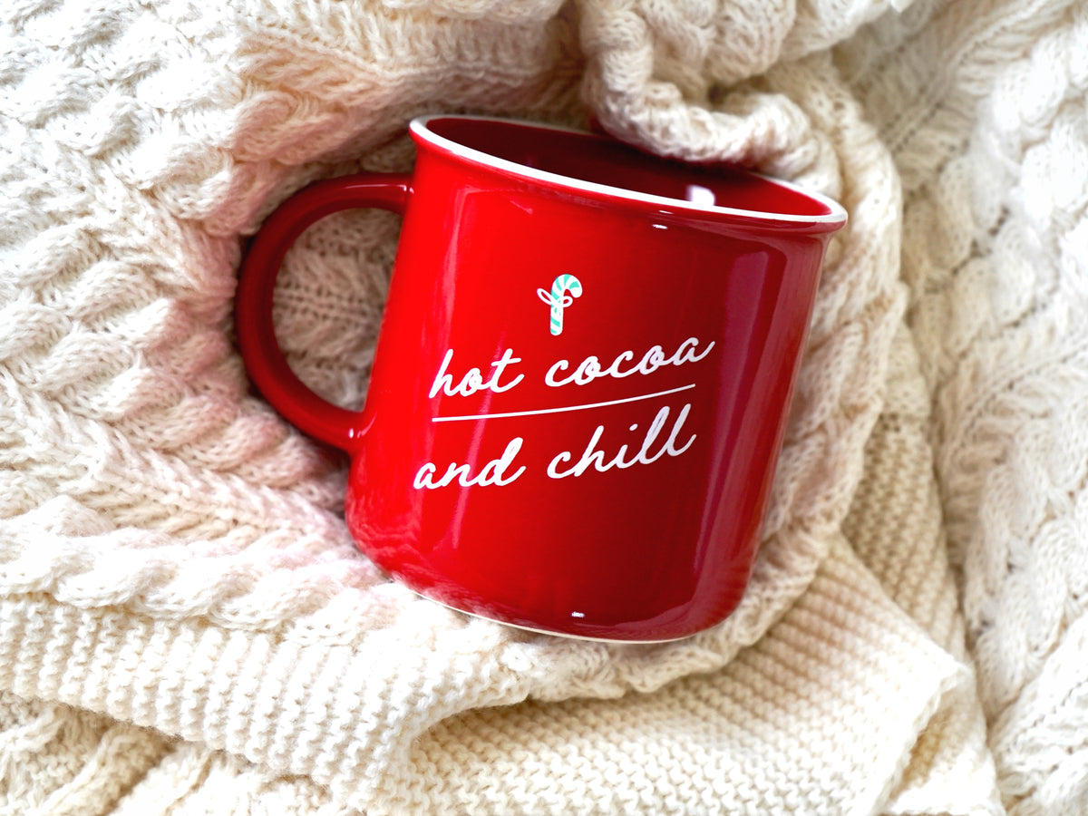 Holiday mug shown is a red ceramic mug with a white rim with the words hot cocoa and chill in script with a mini candy cane 