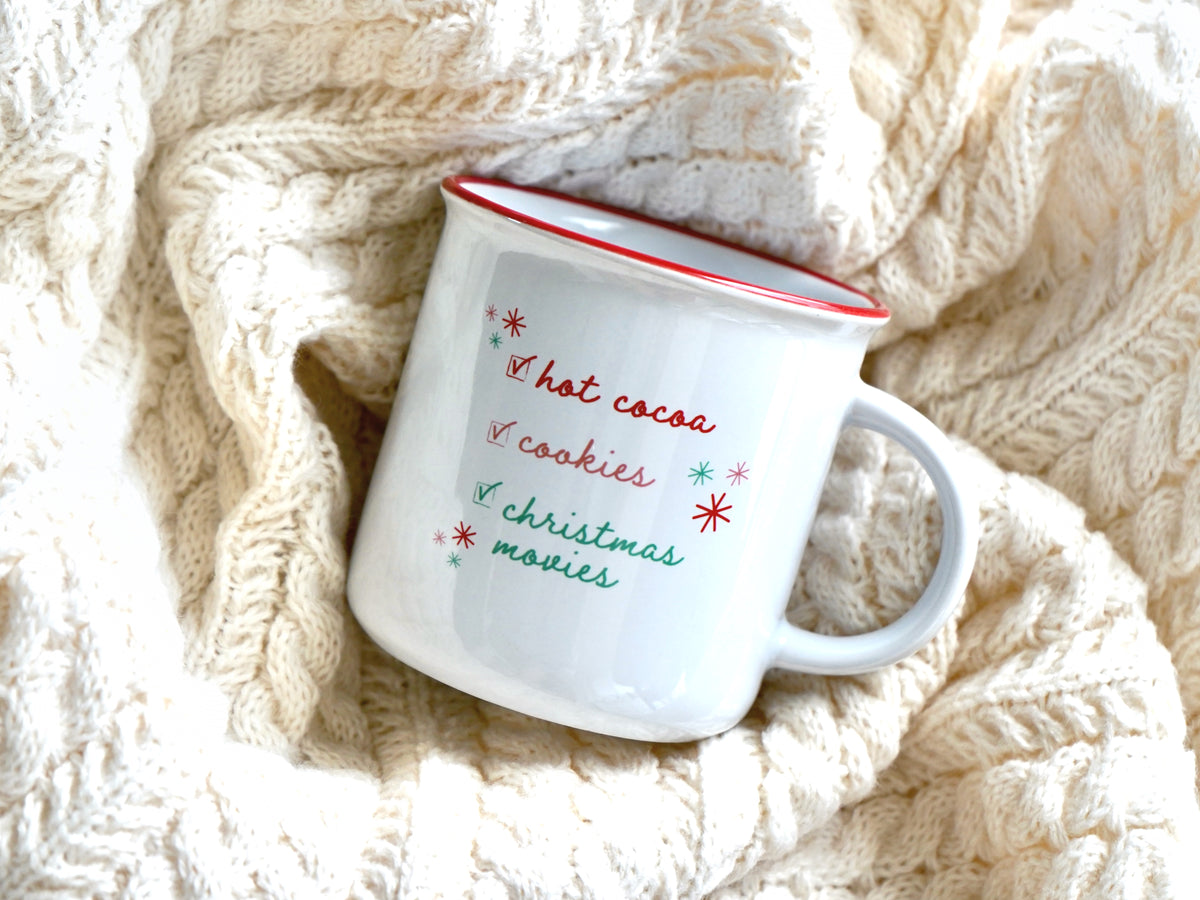Holiday mug is shown in a white ceramic with a red rim with a holiday checklist of hot cocoa, cookies, and Christmas movies in multi colored script font