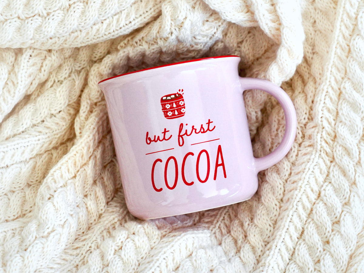 Holiday mug is shown in a light pink with a red rim with the words but first cocoa, and a picture of a hot cocoa 