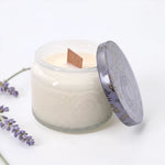 A lavender scented candle in a frosted glass jar with a patterned lid leaning against it, set on a white surface with lavender sprigs in the corner.