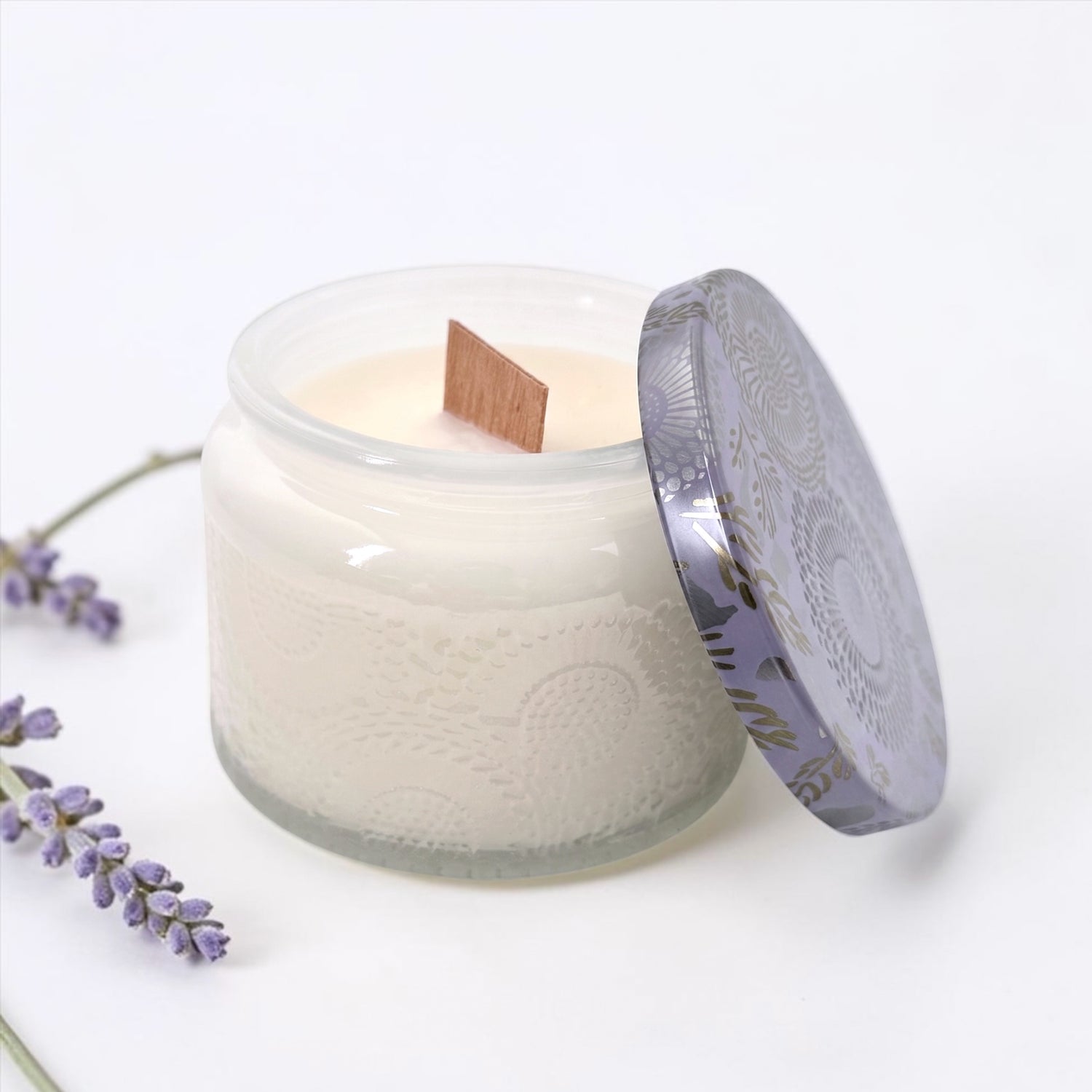 A lavender scented candle in a frosted glass jar with a patterned lid leaning against it, set on a white surface with lavender sprigs in the corner.