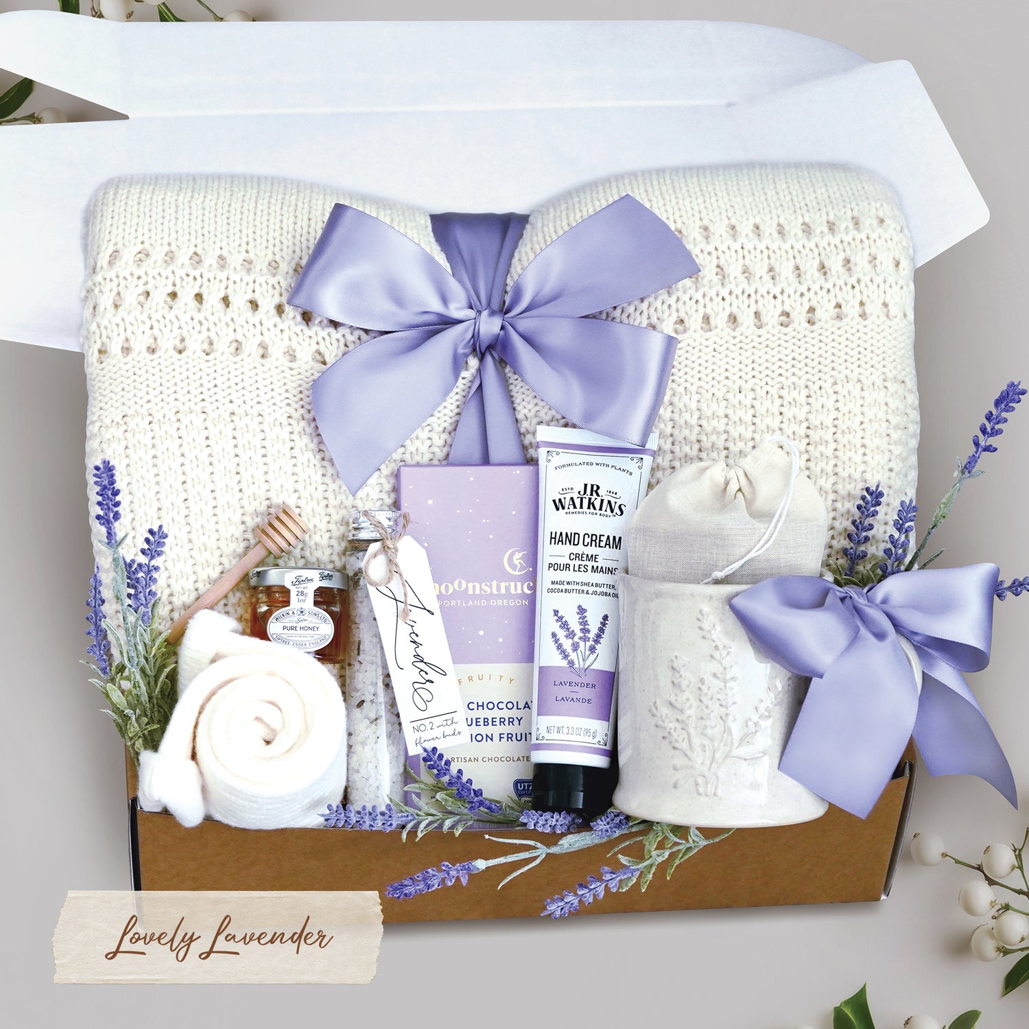 Lovely Lavender comes with a cream lace knit blanket tied with a lavender ribbon, cream knit socks, lavender ceramic mug, 2 tea bags, moonstruck dark chocolate blueberry and passionfruit bar, JR Watkins Lavender hand cream, tube of floral bath salts, mini honey and dipper, and faux lavender sprig