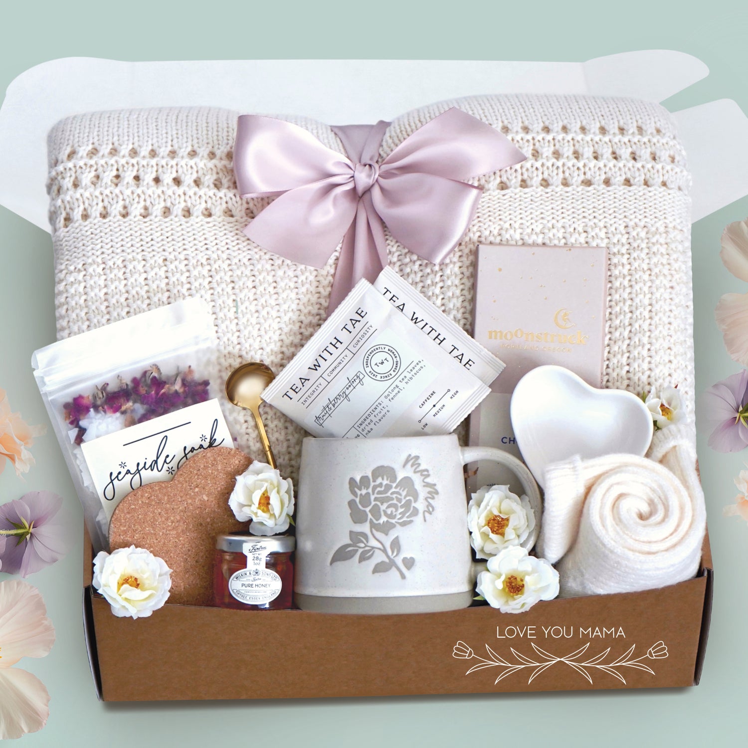 Love you mama blanket box comes with a cream lace knit blanket tied in a blush ribbon, 2 tea with tae tea bags, gold demitasse spoon, rose seaside soak bath salts, heart coaster, mini honey and dipper, mama ceramic mug, heart trinket dish, full size moonstruck milk chocolate bar, and cream knit socks