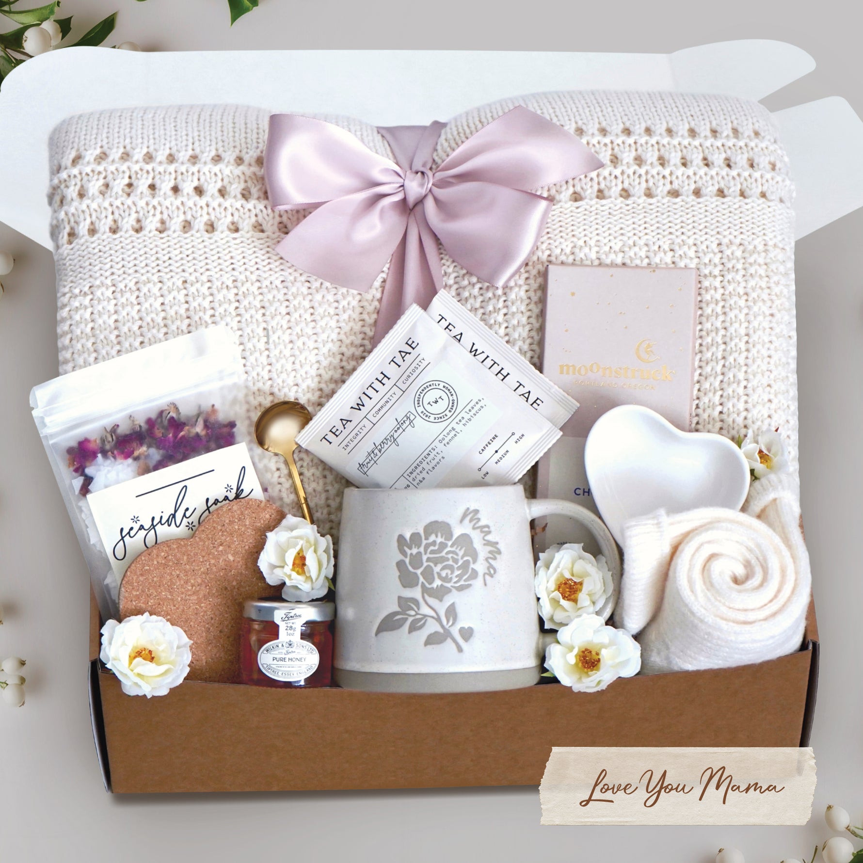Love you mama blanket box comes with a cream lace knit blanket tied in a blush ribbon, 2 tea with tae tea bags, gold demitasse spoon, rose seaside soak bath salts, heart coaster, mini honey, mama ceramic mug, heart trinket dish, full size moonstruck milk chocolate bar, and cream knit socks