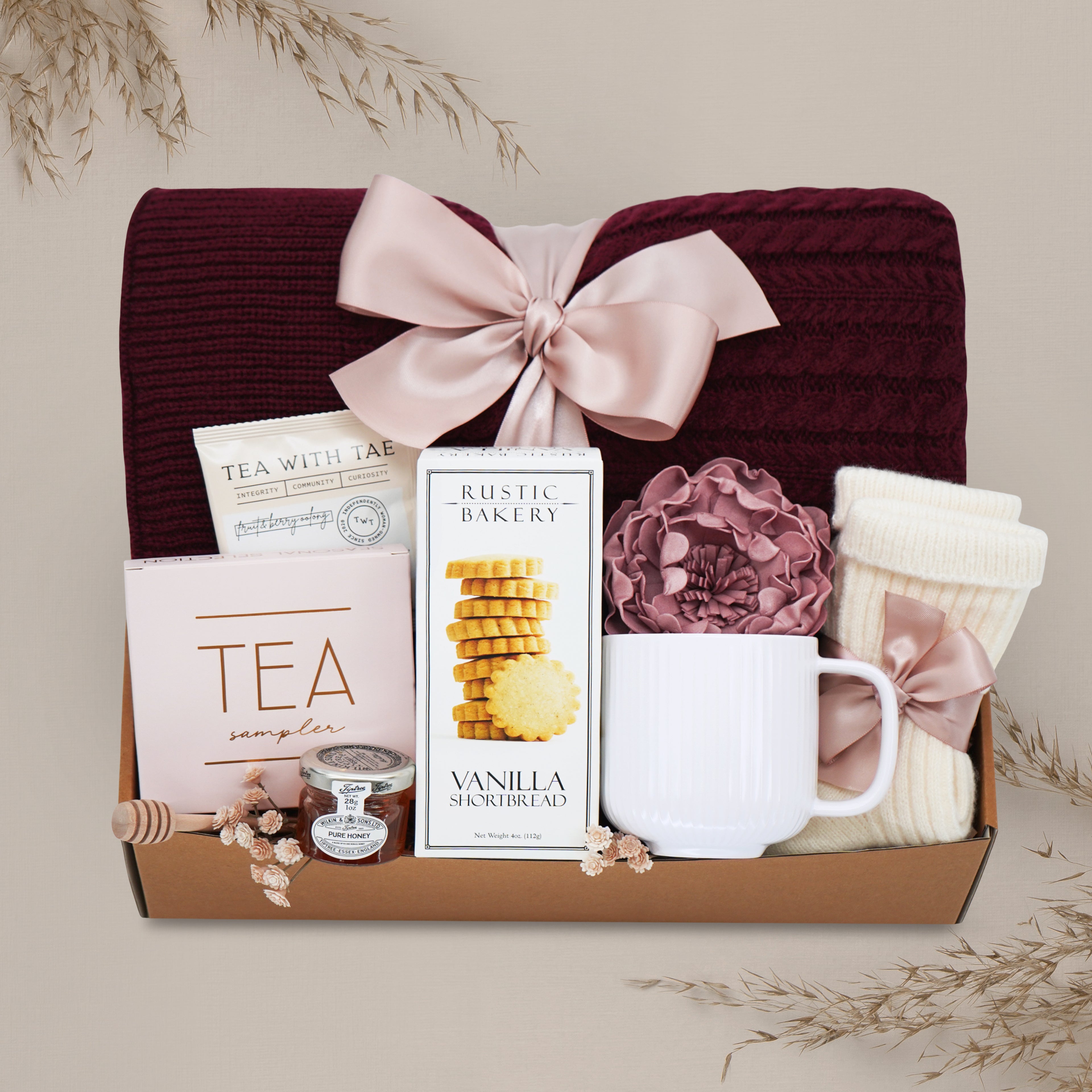 Peaceful Peony by Wildflower Pop comes with a Burgundy cable knit blanket, Rustic Bakery shortbread cookies, Tea Sampler, mini honey and dipper, cream ribbed ceramic mug, cream knit socks, and faux peony