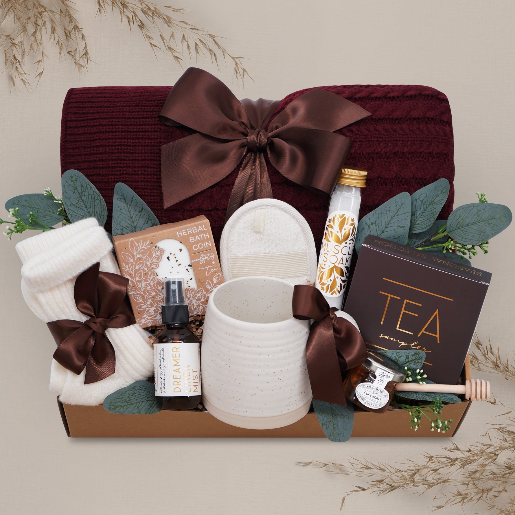 Wildflower Pop Ruby Spa Box with Burgundy blanket, bath salts, bath bomb, linen mist, cream socks, tea sampler, and mini honey and dipper. On tan grey background 