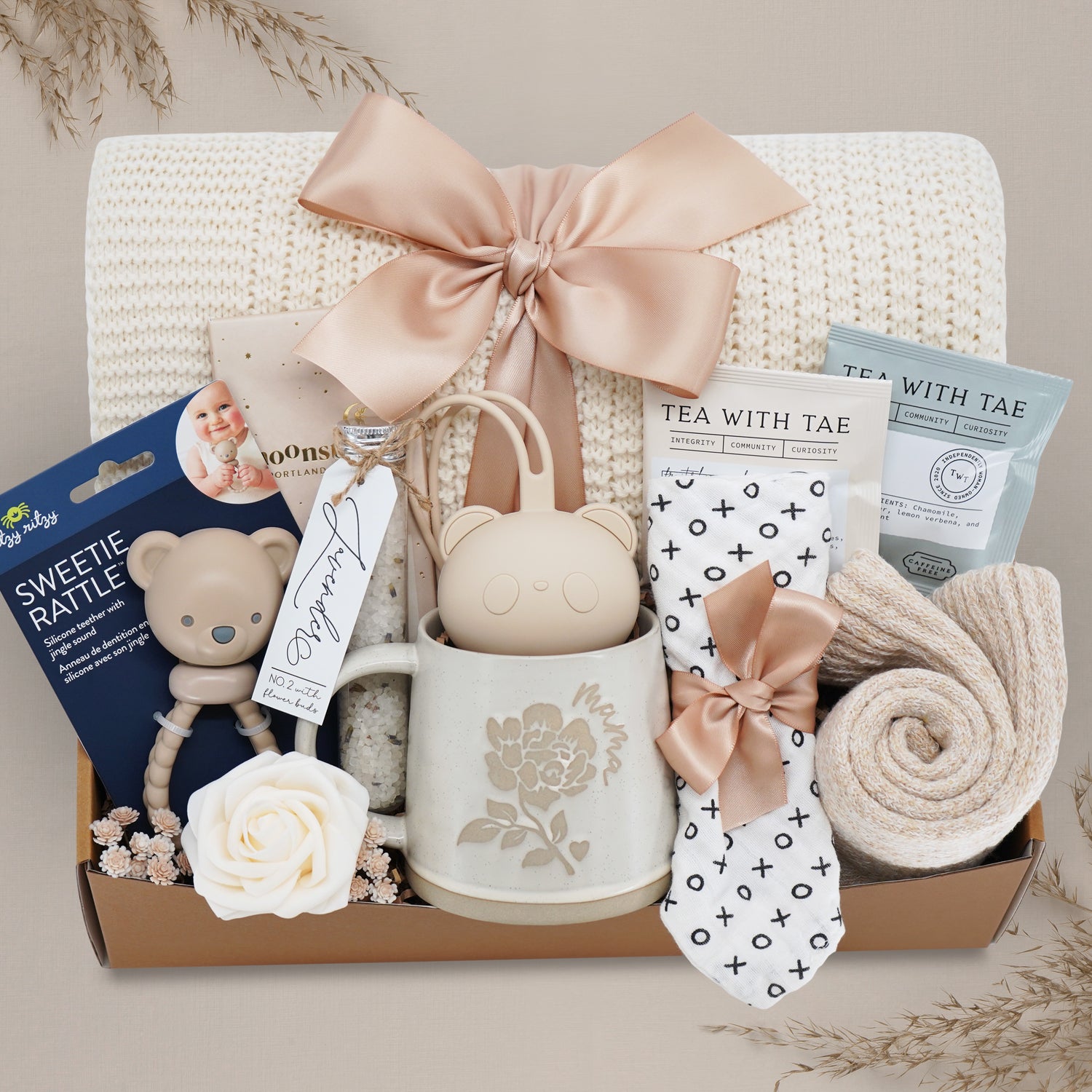 Wildflower Pop Mama Bear Basket comes with a cream lace knit blanket with nude ribbon, Itzy Ritzy Bear Sweetie Rattle, Bear pacifier holder, xo bib, Wildflower Pop Mama mug, floral bath salts, Moonstruck milk chocolate, tea sampler, and cozy boot socks 
