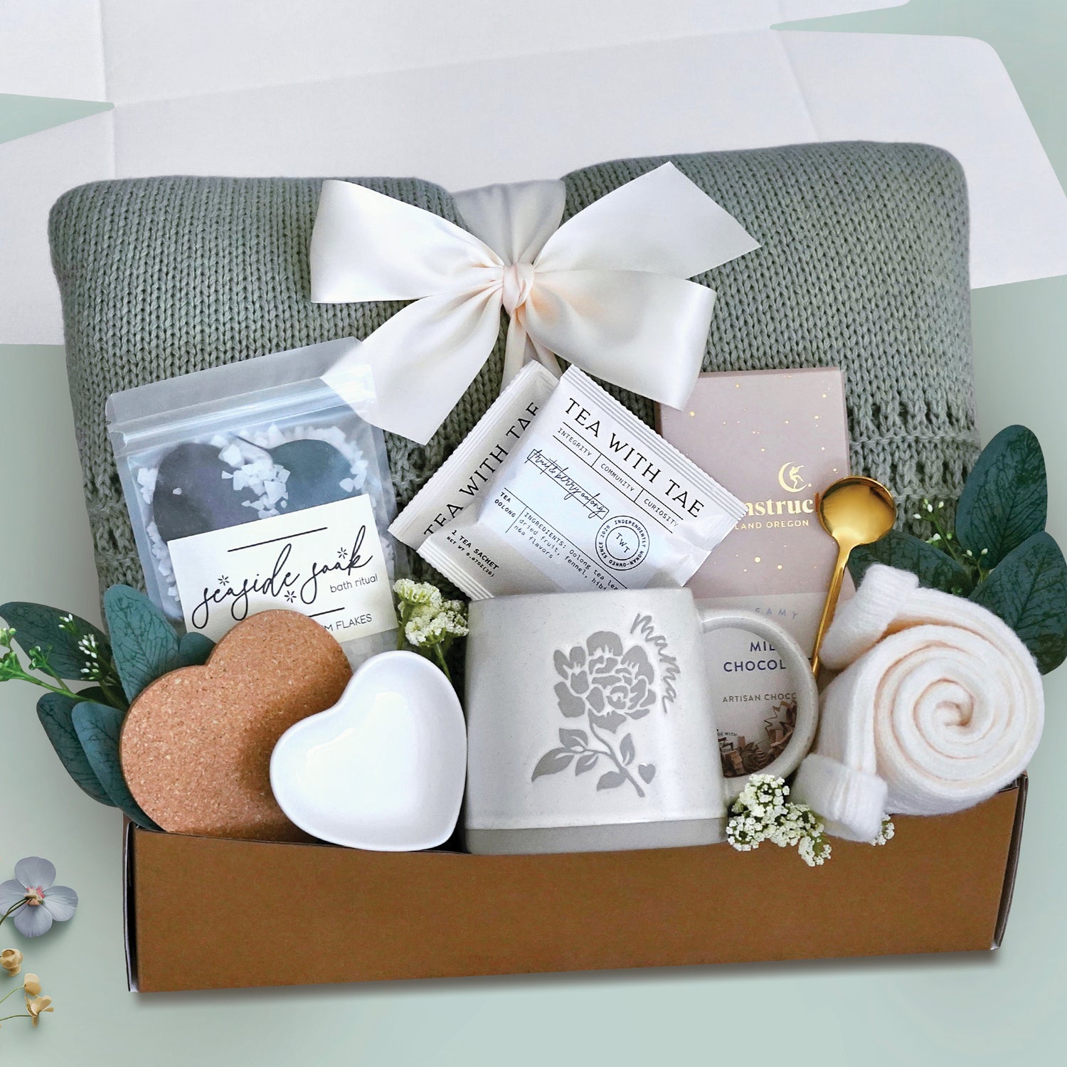 Moms spa retreat blanket box comes with a sage lace knit blanket tied with a cream ribbon, seaside soak bath salts, heart coaster, heart trinket tray. mama ceramic mug, 2 tea with tae tea bags, full size moonstruck milk chocolate bar, cream knit socks, gold demitasse spoon, and faux eucalyptus sprig 