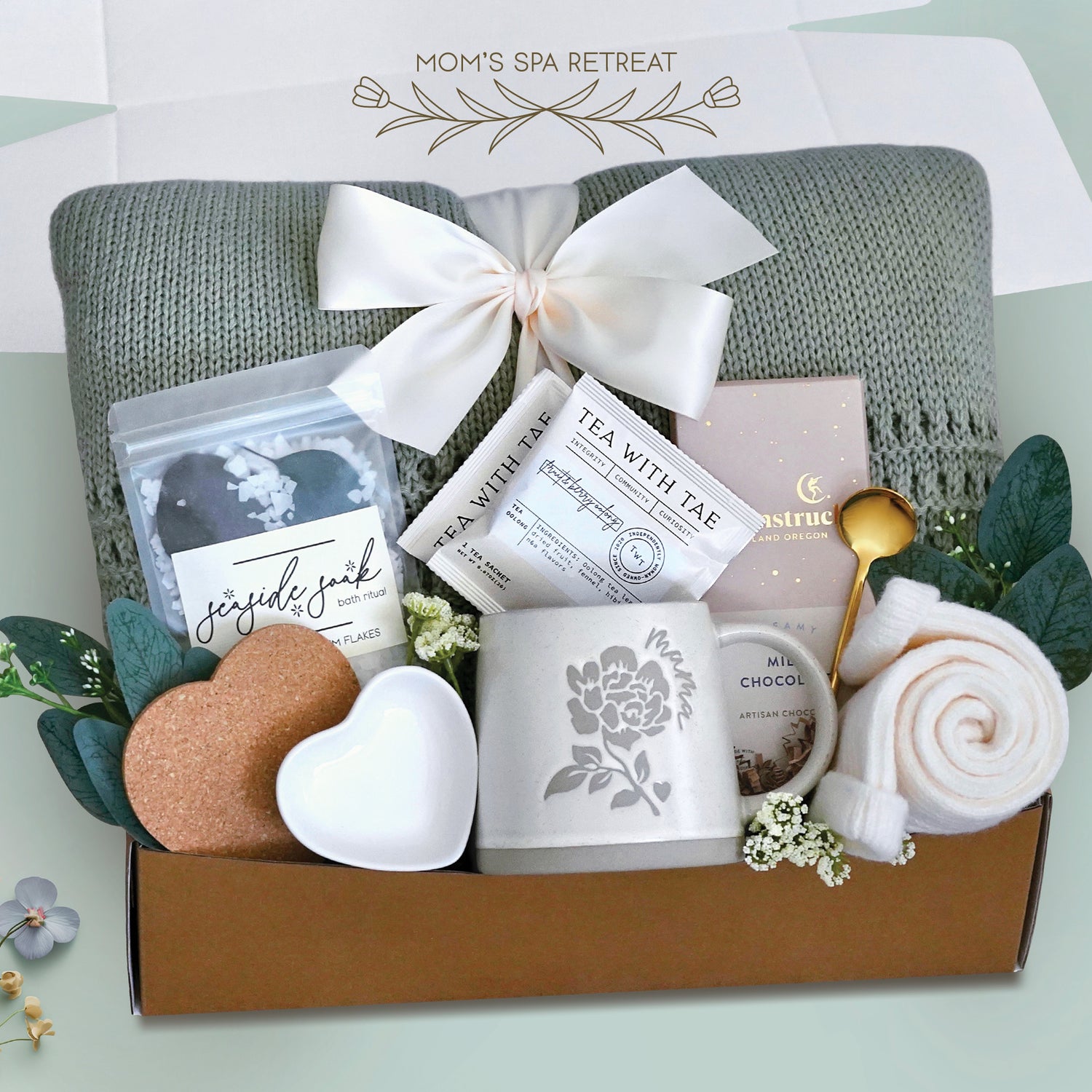 Moms spa retreat blanket box comes with a sage lace knit blanket tied with a cream ribbon, seaside soak bath salts, heart coaster, heart trinket tray. mama ceramic mug, 2 tea with tae tea bags, full size moonstruck milk chocolate bar, cream knit socks, gold demitasse spoon, and faux eucalyptus sprig 