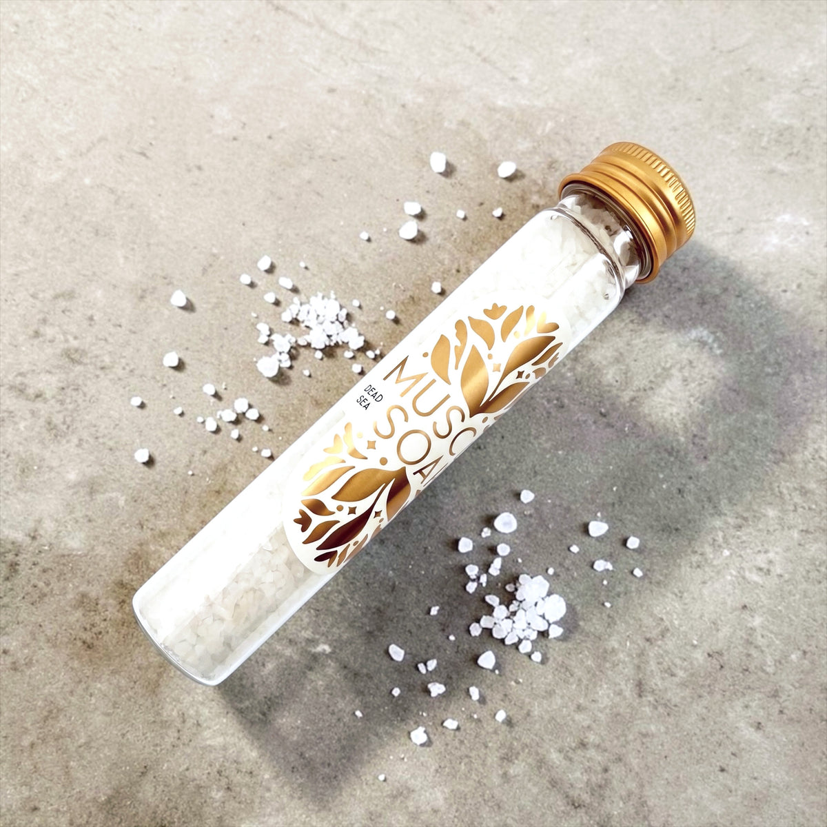 A clear tube with a gold screw cap, labeled 'MUSCLE SOAK' with a gold leaf design, lying diagonally on a textured gray concrete surface.