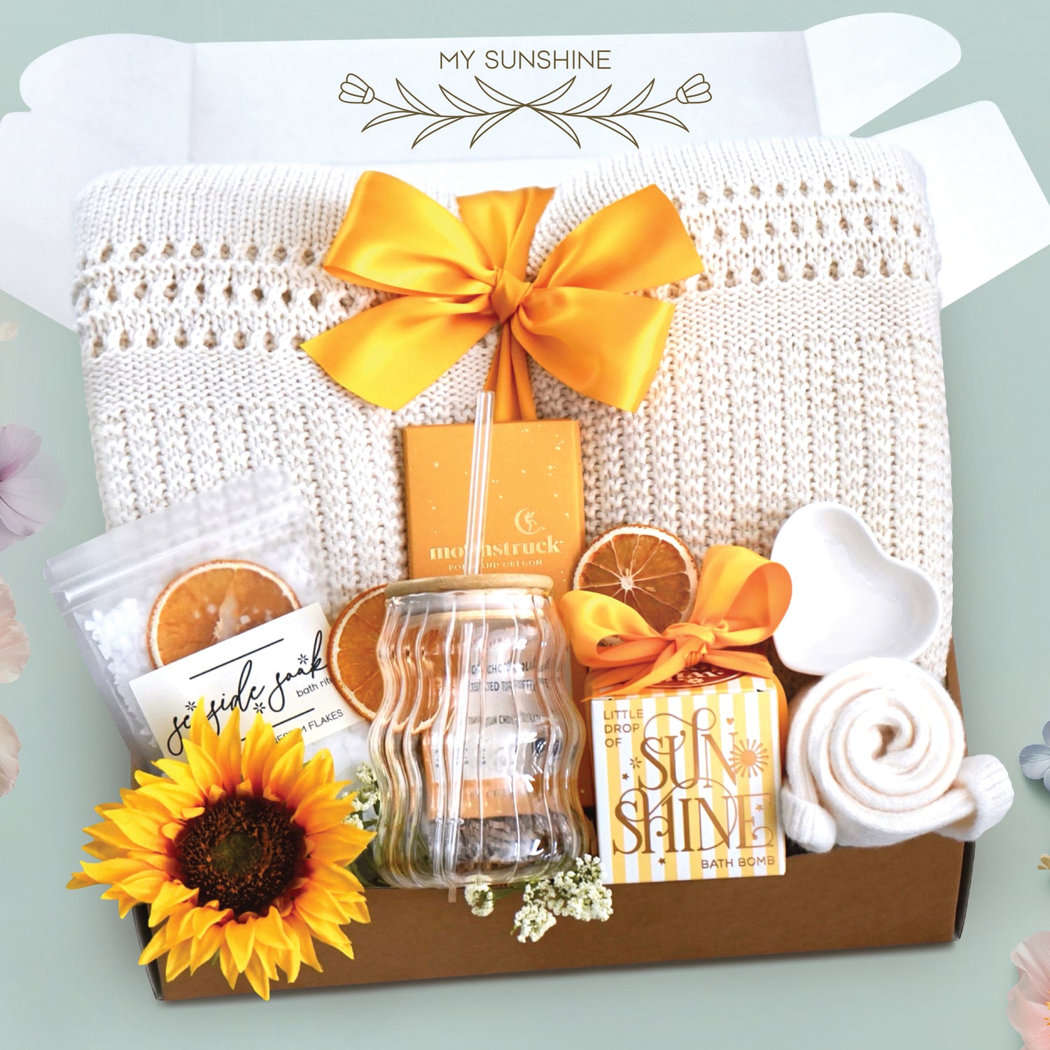 My sunshine blanket box comes with a cream lace knit blanket tied with a yellow ribbon, seaside soak bath salts with orange slices, wavy glass tumbler and straw, full size moonstruck milk chocolate and toffee bar, little drop of sunshine bath bomb, heart trinket tray, and cream knit socks