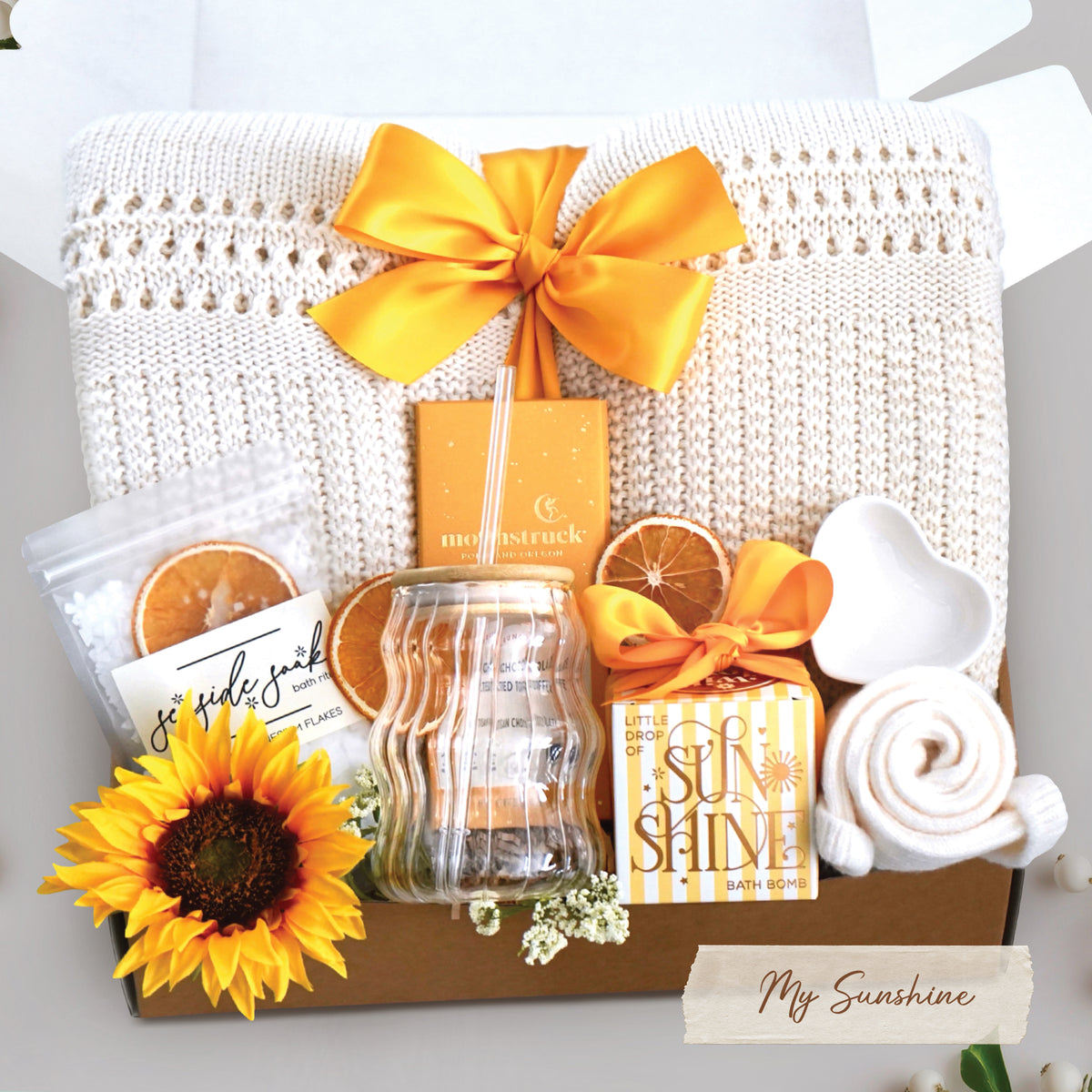 My sunshine blanket box comes with a cream lace knit blanket tied with a yellow ribbon, seaside soak bath salts with orange slices, wavy glass tumbler and straw, full size moonstruck milk chocolate and toffee bar, little drop of sunshine bath bomb, heart trinket tray, and cream knit socks