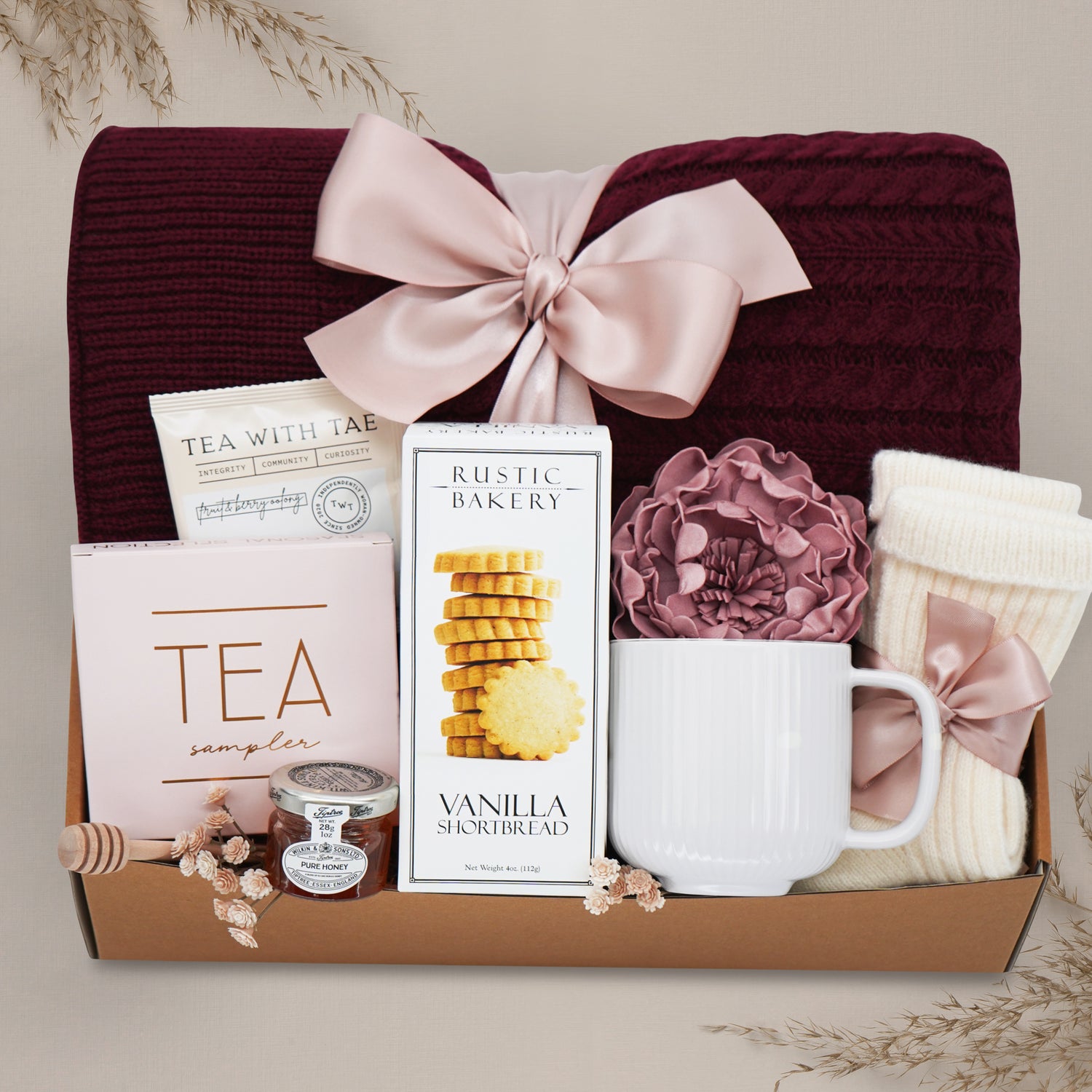 Peaceful Peony by Wildflower Pop comes with a Burgundy cable knit blanket, Rustic Bakery shortbread cookies, Tea Sampler, mini honey and dipper, cream ribbed ceramic mug, cream knit socks, and faux peony