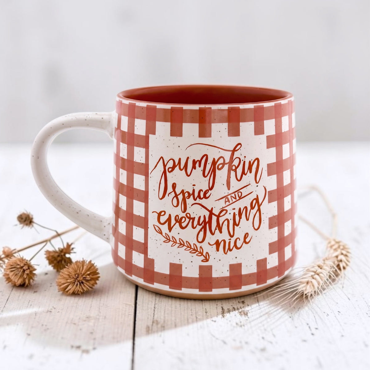 A ceramic mug with a white and terracotta gingham pattern, featuring the phrase 'pumpkin spice and everything nice' in script font. The mug is styled on a white wooden surface with dried flowers and wheat stalks.