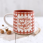A ceramic mug with a white and terracotta gingham pattern, featuring the phrase 'pumpkin spice and everything nice' in script font. The mug is styled on a white wooden surface with dried flowers and wheat stalks.