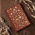 A Wildflower Pop hardcover notebook with a deep reddish-brown cover decorated with gold foil autumn illustrations of pumpkins, leaves, and mushrooms, displayed on a dark wood background with dried wheat sprigs.