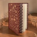 A Wildflower Pop hardcover journal standing upright on a wooden table, featuring a dark red cover with gold-toned autumn illustrations like pumpkins, leaves, and acorns, plus a dark gingham patterned spine and white pages.