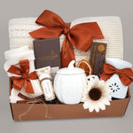 Wildflower Pop Pumpkin Spice Blanket Gift Box, cream cable knit blanket with pumpkin orange ribbon, chocolate, cream socks, cream pumpkin mug with lid, hand lotion, wax warmer and wax melt. On a grey background