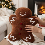 Gingerbread man pillow with white icing details and red buttons, being held by hands in a cozy living room setting with a fireplace and Christmas tree in the background.