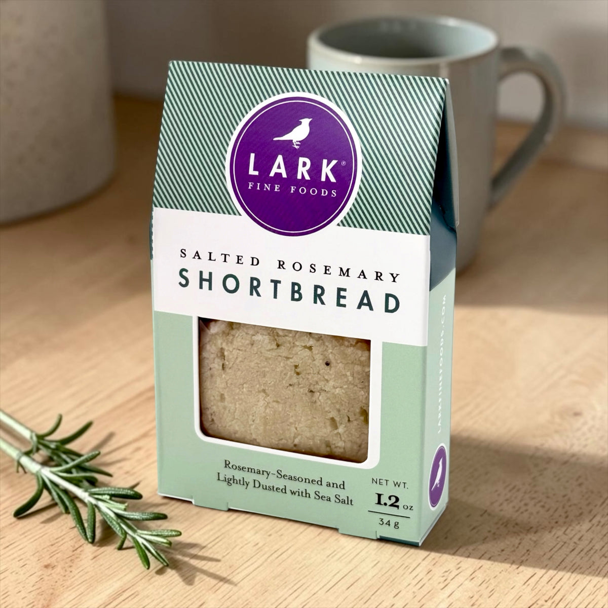 Lark Fine Foods Salted Rosemary Shortbread sits on a wooden table with a sprig of rosemary and a gray mug in the background.