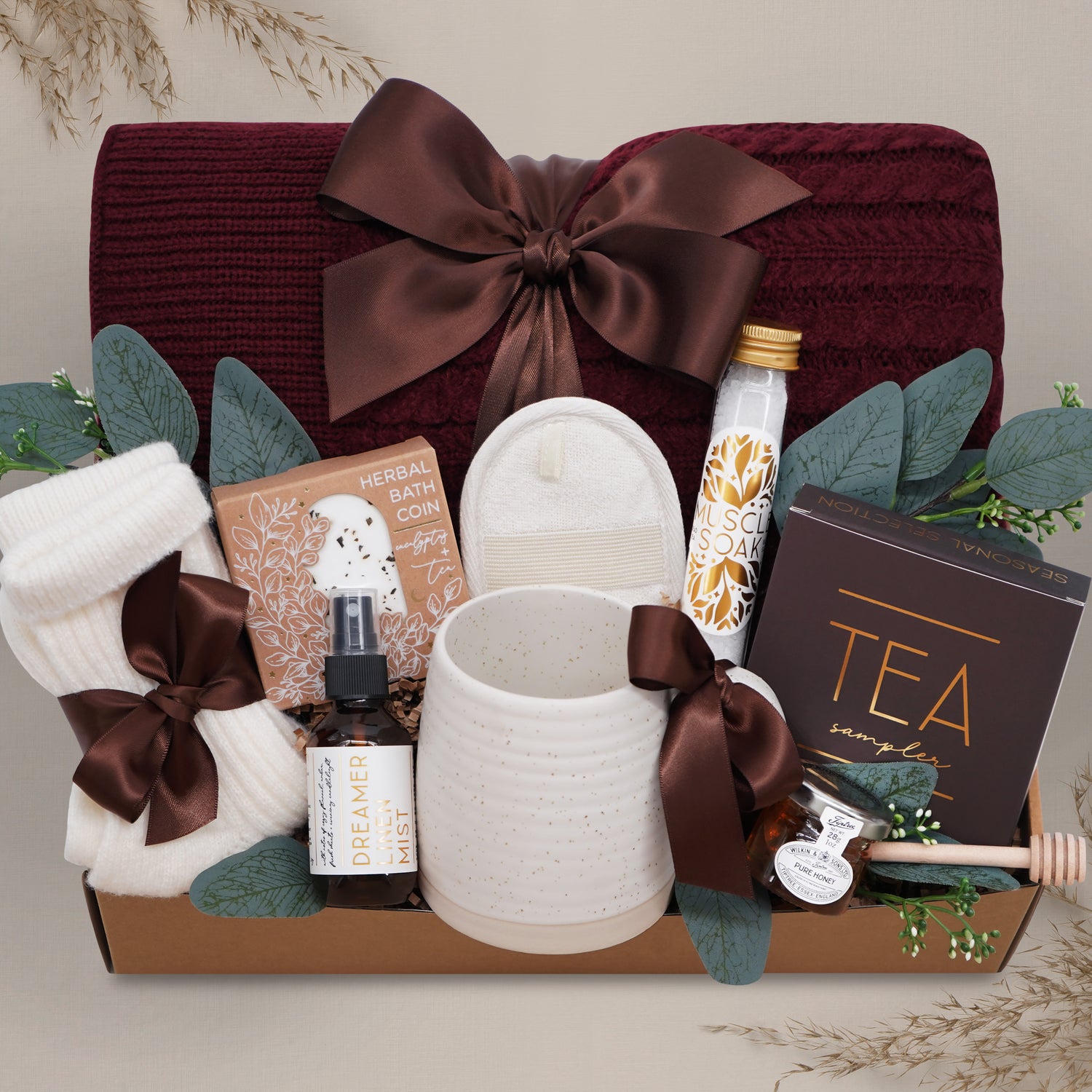 Wildflower Pop Ruby Spa Box with Burgundy blanket, bath salts, bath bomb, linen mist, cream socks, tea sampler, and mini honey and dipper. On tan grey background 