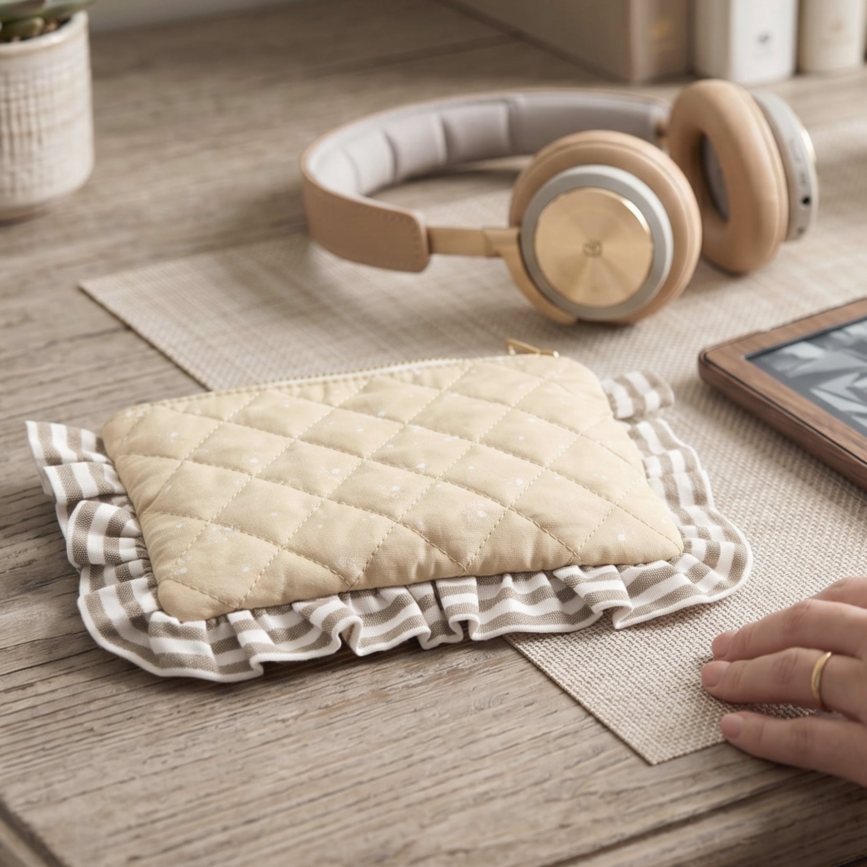 A quilted beige pouch with a striped ruffled border sits on a wooden desk next to headphones and a tablet.
