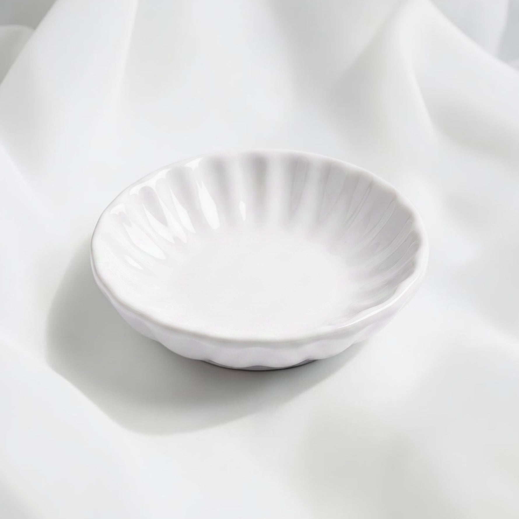 A small white ceramic trinket dish with a fluted, scalloped rim, photographed on draped white fabric.