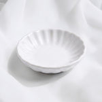 A small white ceramic trinket dish with a fluted, scalloped rim, photographed on draped white fabric.