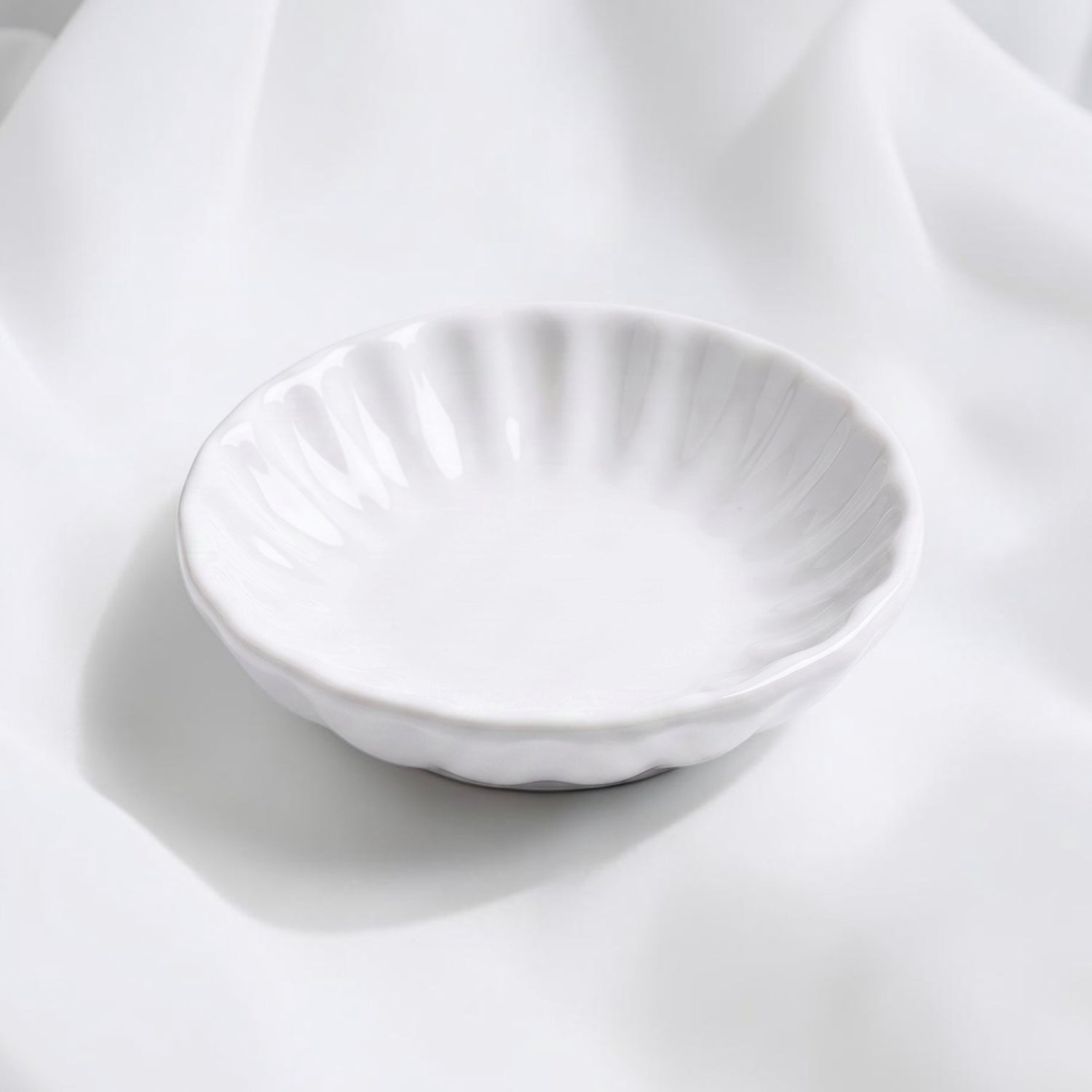 A small white ceramic trinket dish with a fluted, scalloped rim, photographed on draped white fabric.