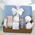 The Sea Glass Blanket Box comes with a blue lace knit blanket tied in a cream ribbon, full size Moonstruck milk chocolate bar, White tea bath salts, Old Whaling co floral coast bath bomb, scallop trinket dish, wave glass tumbler with plastic straw, sea shell ornament, and cream knit socks 