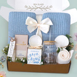 The Sea Glass Blanket Box comes with a blue lace knit blanket tied in a cream ribbon, full size Moonstruck milk chocolate bar, White tea bath salts, Old Whaling co floral coast bath bomb, scallop trinket dish, wave glass tumbler with plastic straw, sea shell ornament, and cream knit socks 