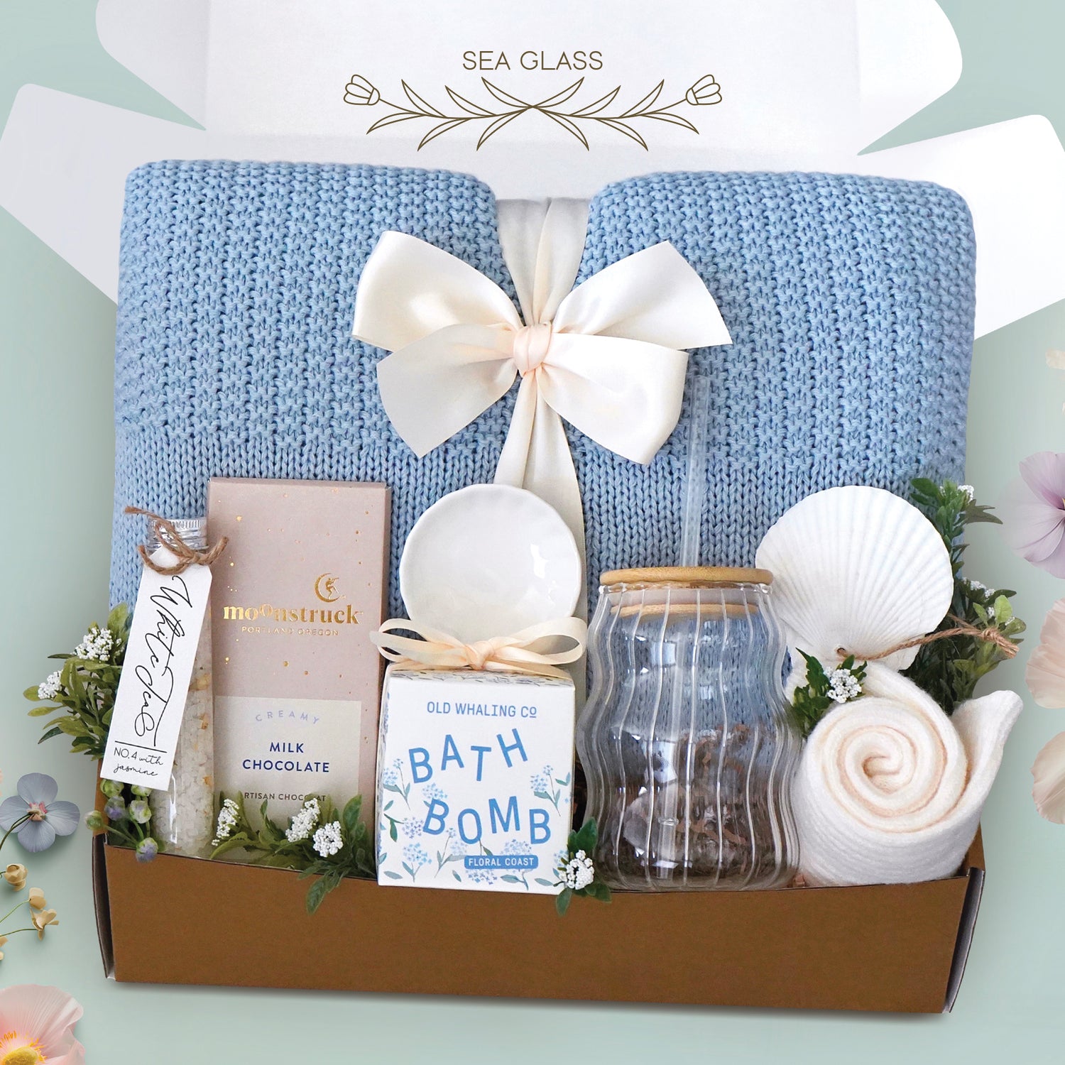 The Sea Glass Blanket Box comes with a blue lace knit blanket tied in a cream ribbon, full size Moonstruck milk chocolate bar, White tea bath salts, Old Whaling co floral coast bath bomb, scallop trinket dish, wave glass tumbler with plastic straw, sea shell ornament, and cream knit socks 