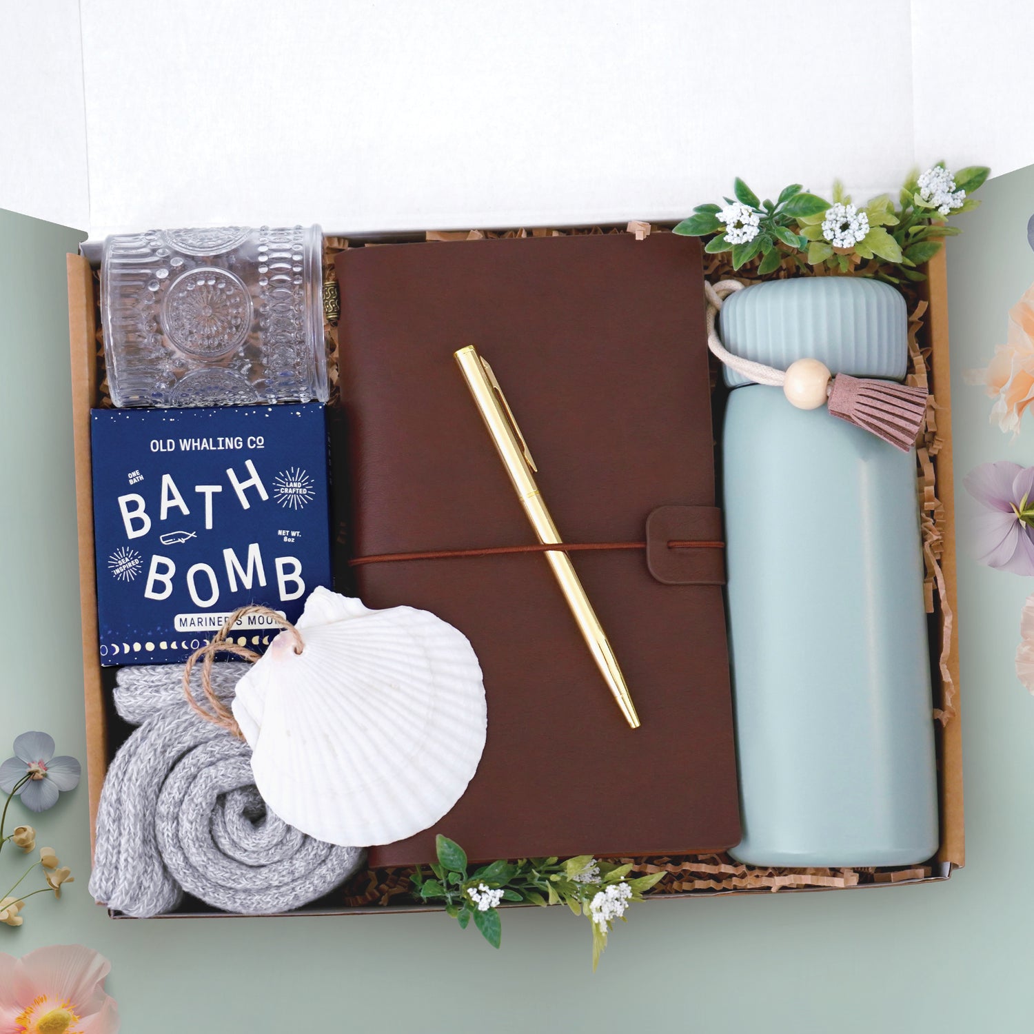 The seaside memories tumbler box comes with a sea foam green insulated tumbler, brown leatherette travelers notebook, gold ball point pen, mariners moon bath bomb, glass votive and tea light, cozy boot socks, and seashell ornament 