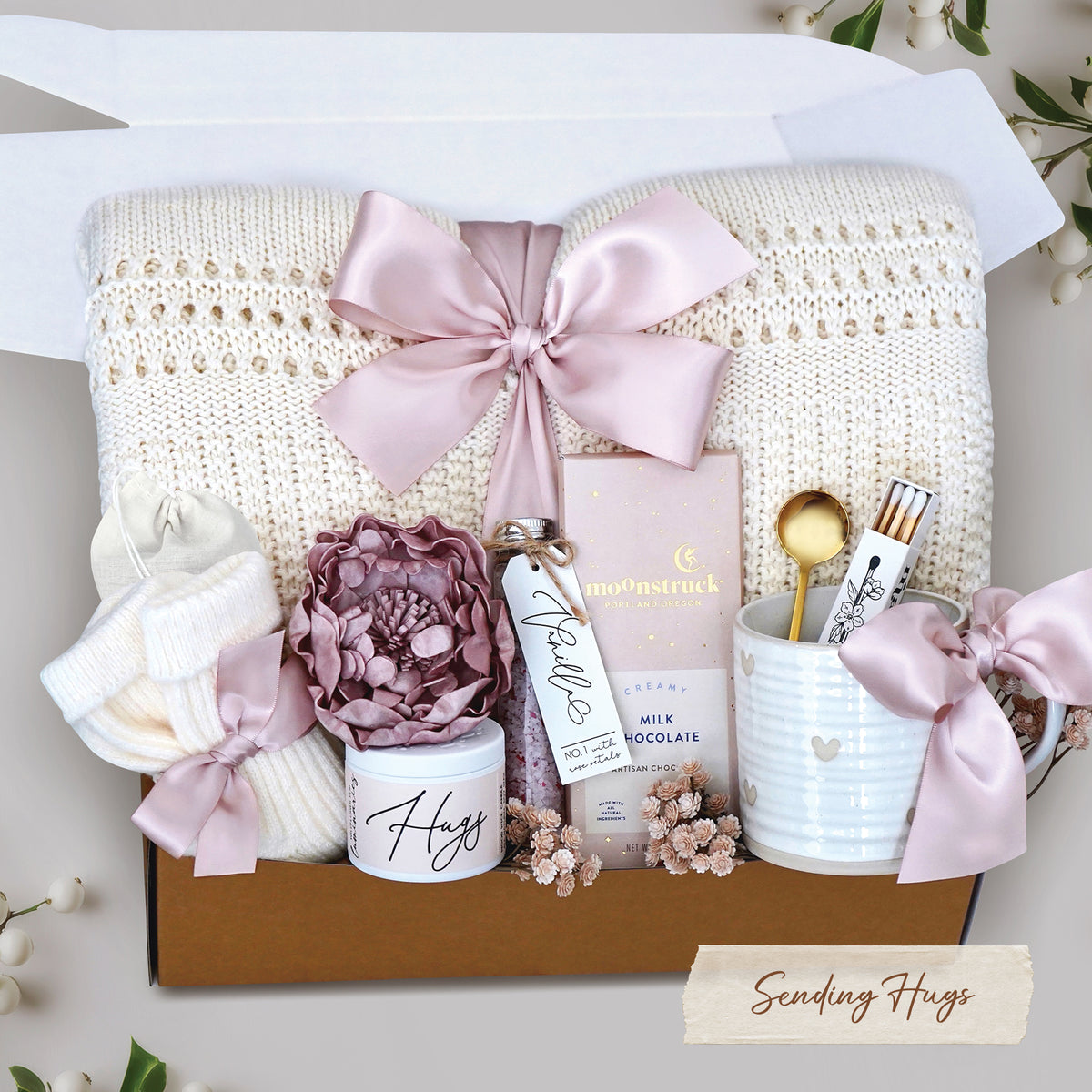 Sending Hugs Gift Box with a cream lace knit blanket, heart ceramic mug, matches, bath salt tube, faux peony head, cream knit socks, moonstruck milk chocolate bar. Finished with blush ribbon and packaged in a branded Wildflower Pop Gift Box