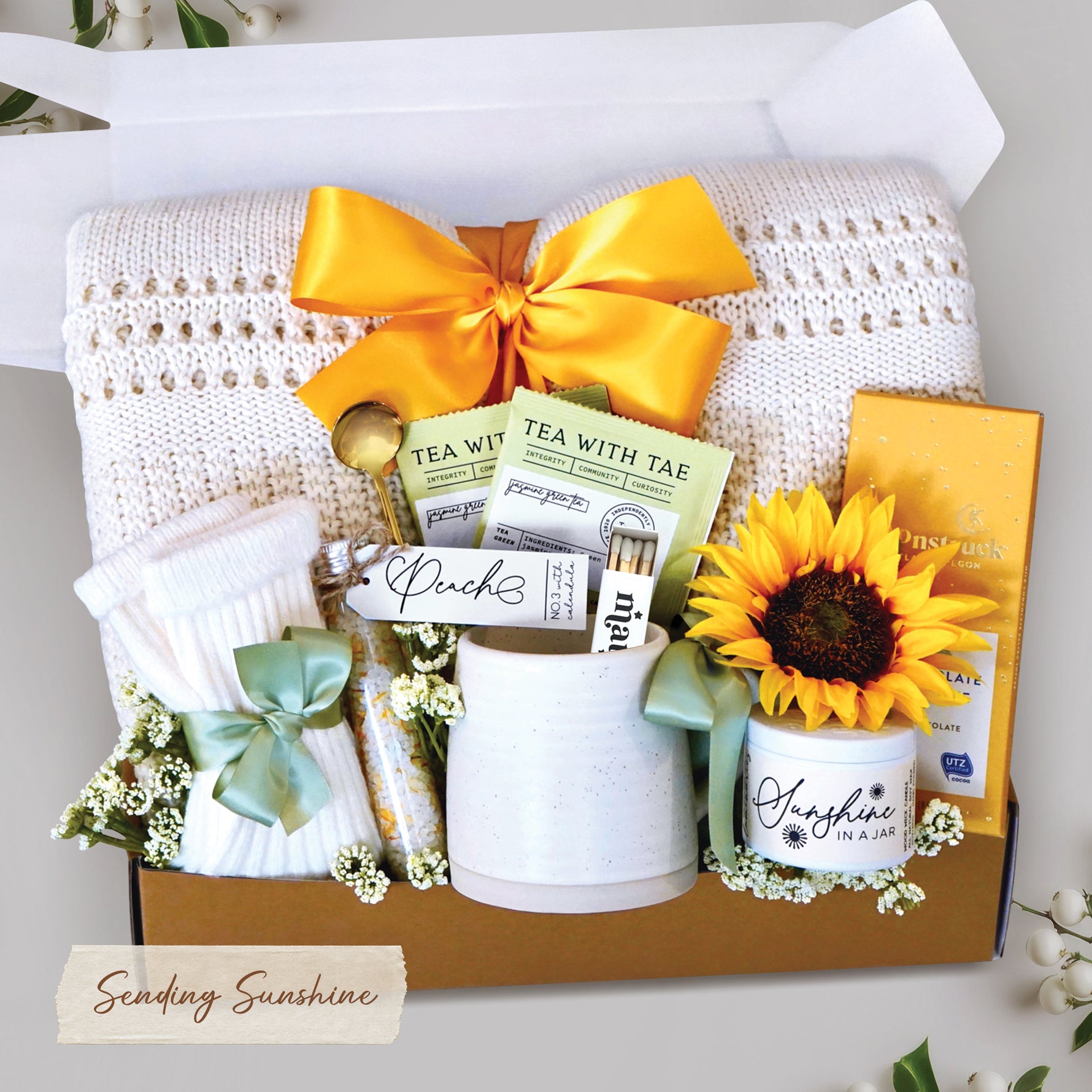 Sending Sunshine Gift Box comes with cream lace knit throw tied with sunshine yellow ribbon, tea sampler, tube of bath salts, trail size soy wax candle and matches, toffee and milk chocolate moonstruck bar, cream knit socks, and faux sunflower head. 