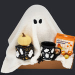 Halloween-themed gift set with ghost pillow, pumpkin, Wildflower Pop Ghost mug, and candy on a dark background.