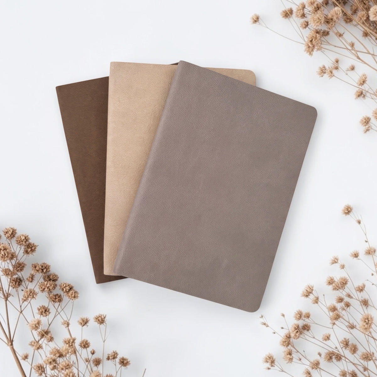 Three faux leather notebooks in brown, beige, and taupe, arranged on a clean white background.