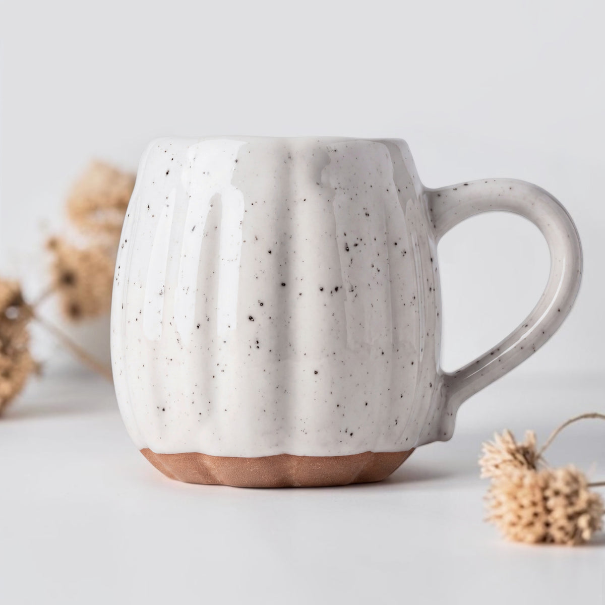 A ceramic mug with a ribbed pumpkin-like design, speckled off-white glaze, and a terracotta-colored base, styled with dried flowers on a light gray background.