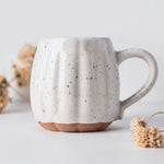 A ceramic mug with a ribbed pumpkin-like design, speckled off-white glaze, and a terracotta-colored base, styled with dried flowers on a light gray background.