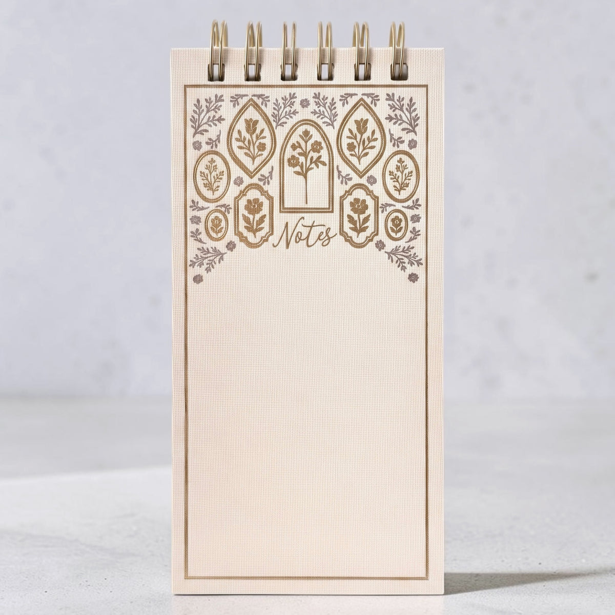 A cream-colored spiral-bound notepad with a decorative floral border in gold and silver foiling. The word 'Notes' are on the cover, shown on a gray background.