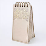 A cream-colored spiral-bound notepad with a decorative floral border in gold and silver foiling. The word 'Notes' are on the cover, shown on a gray background.