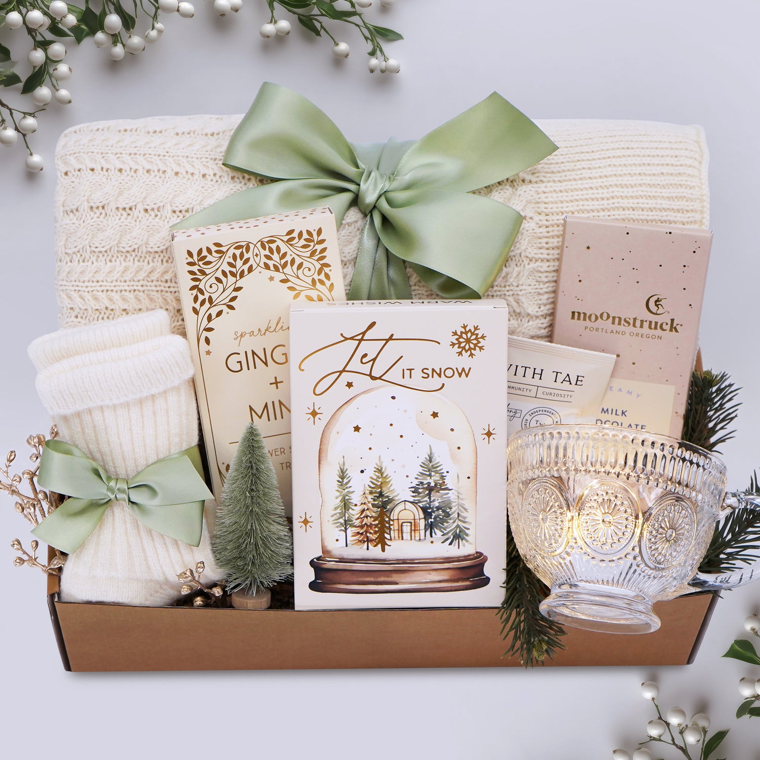 Gift box with decorative items including a candle, card, and small tree, on a light background.