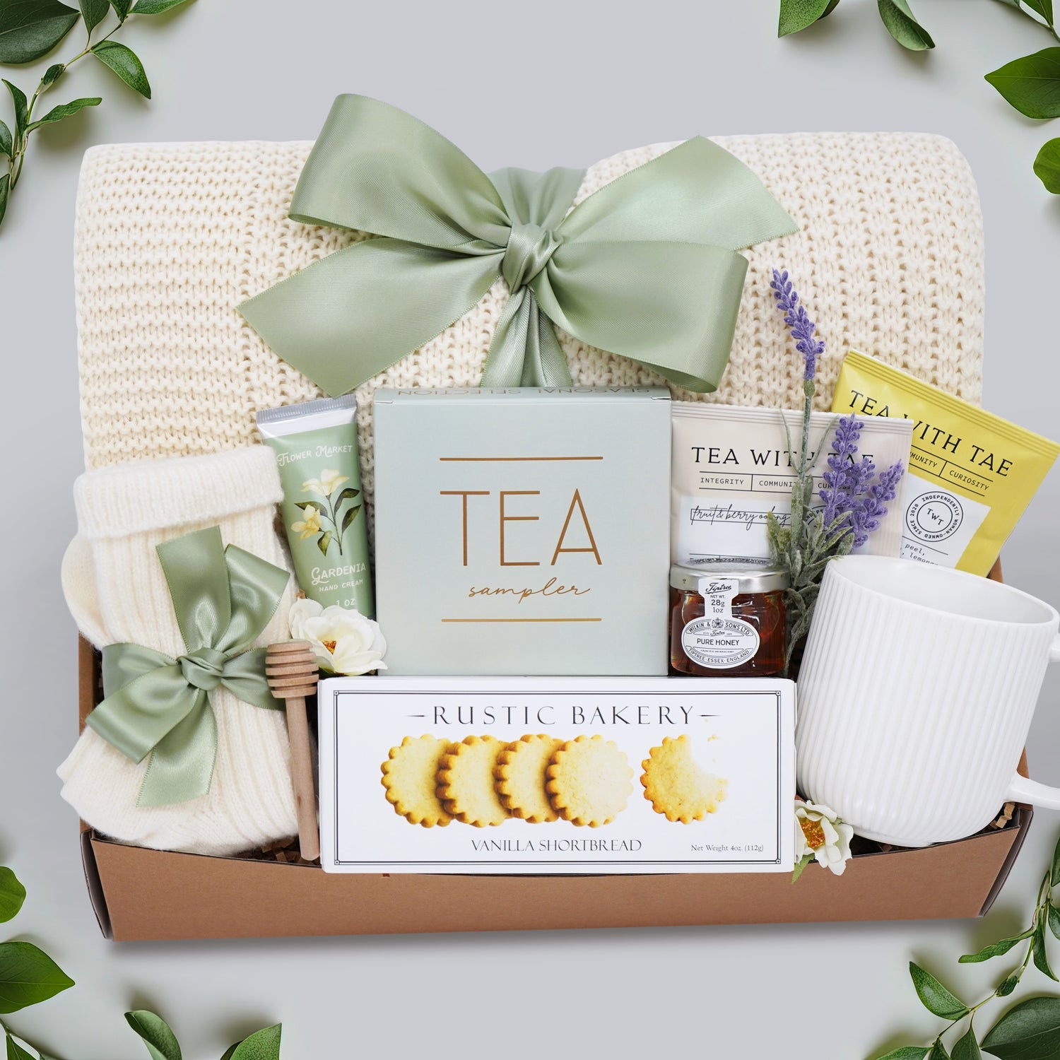Wildflower Pop Tea Party for One comes with a cream lace knit blanket, cream knit socks, Flower Market Gardenia hand cream, Tea Sampler, Rustic Bakery shortbread cookies, ribbed white ceramic mug, mini honey and dipper