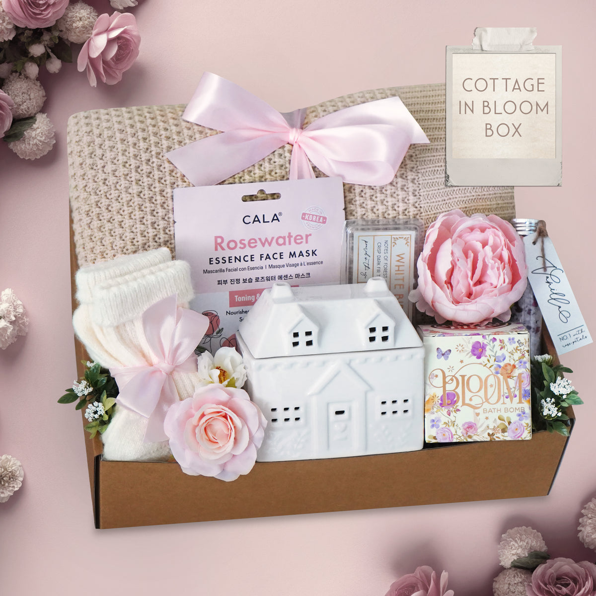 Gift box with decorative items including a blanket, socks, bath salts rosewater face mask, small house-shaped wax warmer, and pink flowers on a pink background.