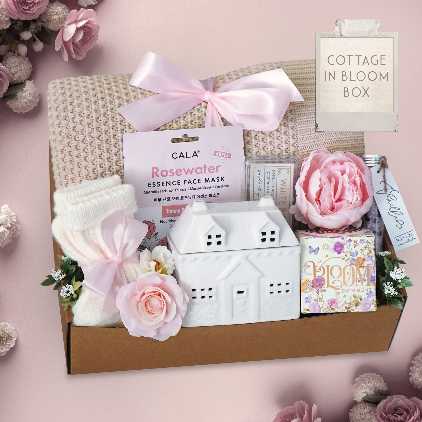 Gift box with decorative items including a blanket, socks, bath salts rosewater face mask, small house-shaped wax warmer, and pink flowers on a pink background.