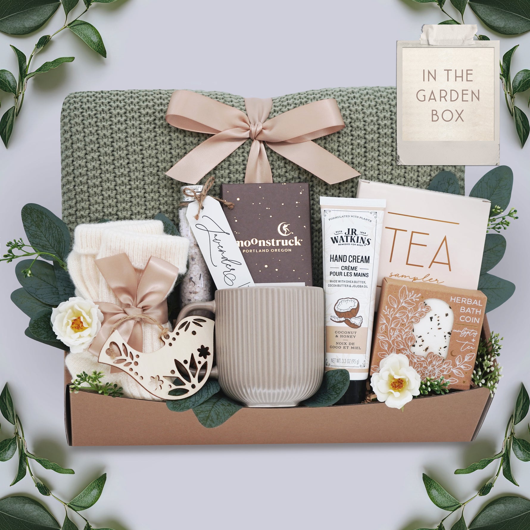 In the Garden Blanket Gift box by Wildflower Pop comes with sage green lace knit blanket, cream knit socks, chocolate bar, floral bath salt tube, grey ribbed mug, hand cream, herbal bath coin, and tea sampler